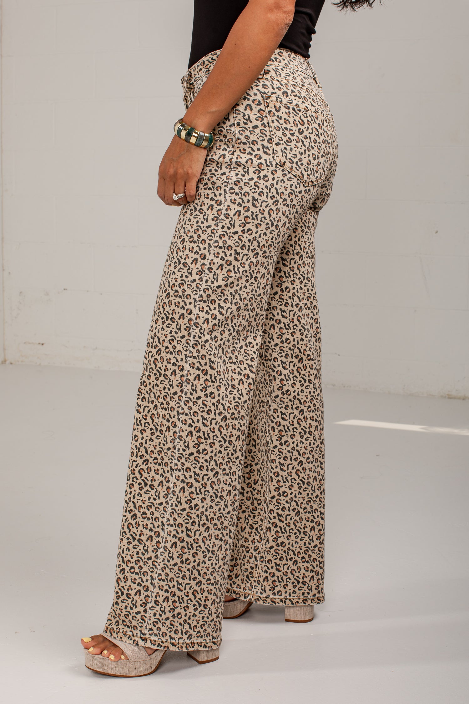 A person wearing high-waisted, wide-leg pants with an allover leopard print pattern in beige, brown, and black. The photo is taken from a side/back angle against a plain white studio background.