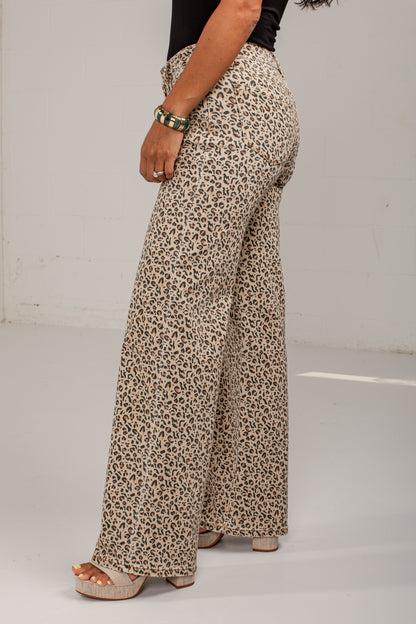 A person wearing high-waisted, wide-leg pants with an allover leopard print pattern in beige, brown, and black. The photo is taken from a side/back angle against a plain white studio background.