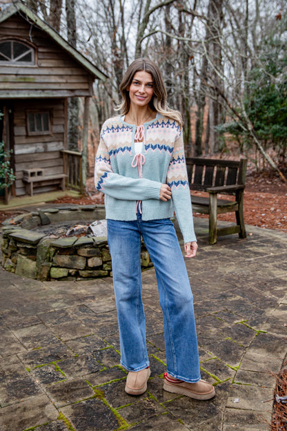 Woman wearing a patterned sweater and jeans standing outdoors near a wooden cabin.