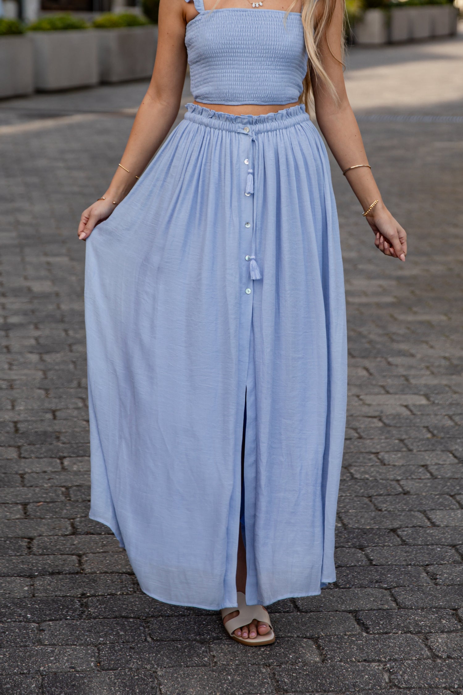 Woman wearing a light blue dress with a button front and wide leg on a paved street.