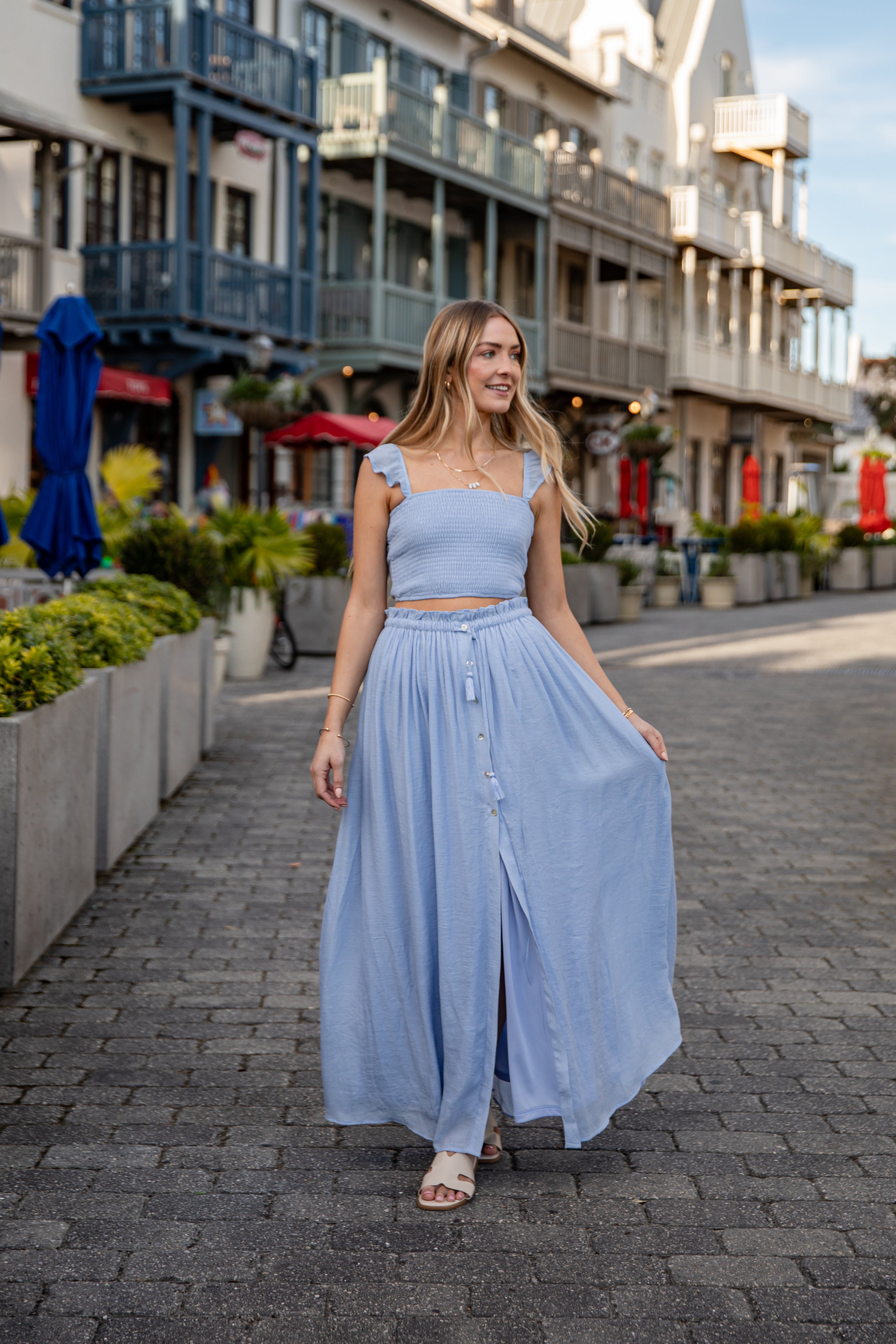 Woman in a light blue outfit walking on a street with shops and plants in the background