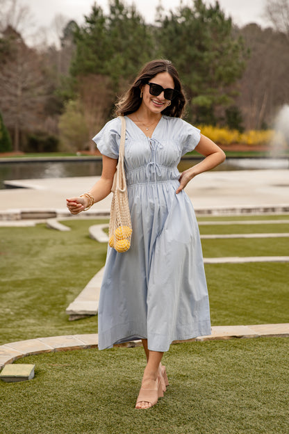 Woman in a light blue dress standing outdoors with a fountain and greenery in the background
