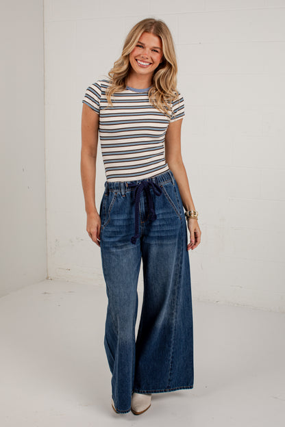 A woman modeling a short-sleeve striped t-shirt and high-waisted wide-leg blue jeans with a drawstring waist, standing against a white wall.