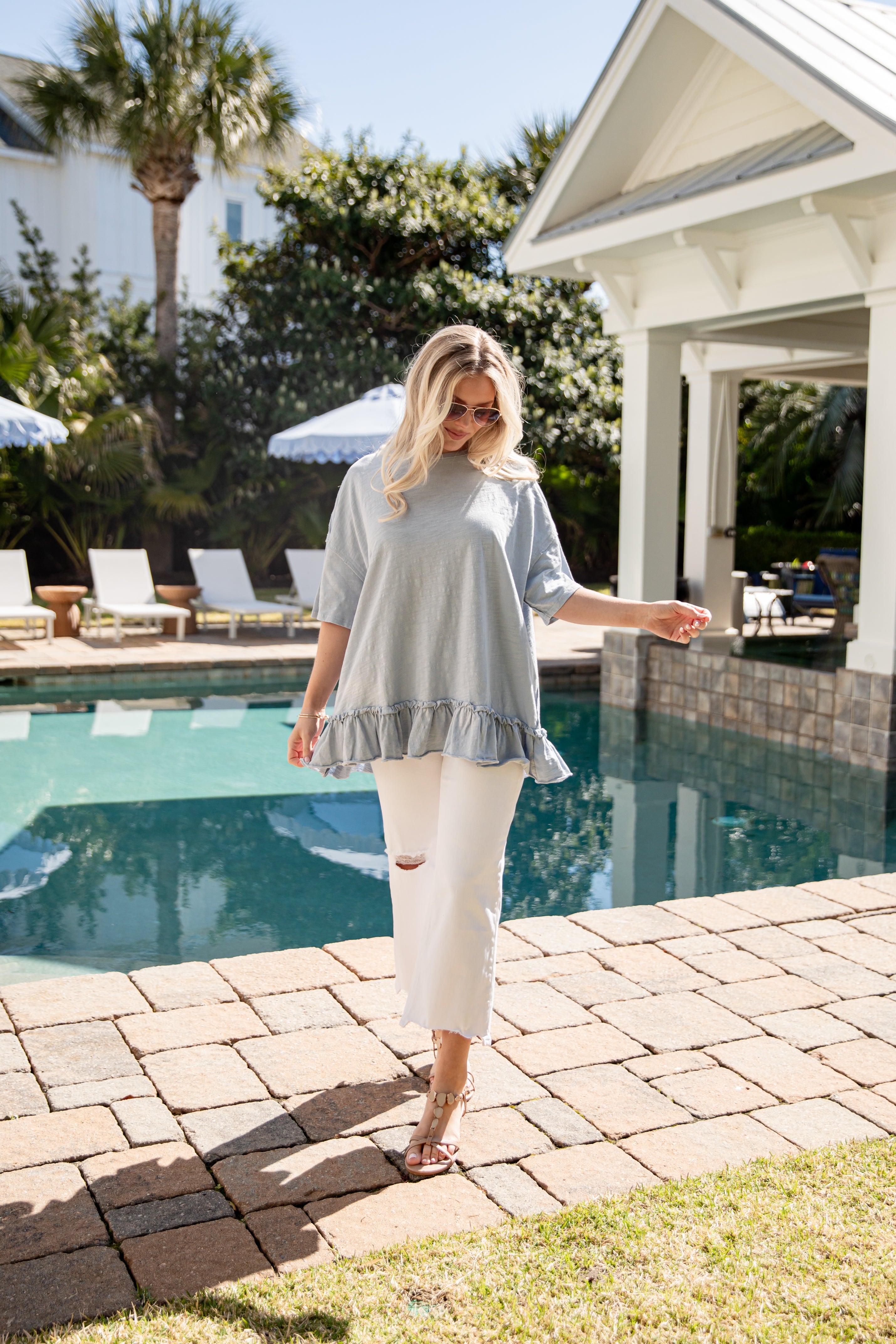 Woman in a light blue top and white pants standing by a poolside.
