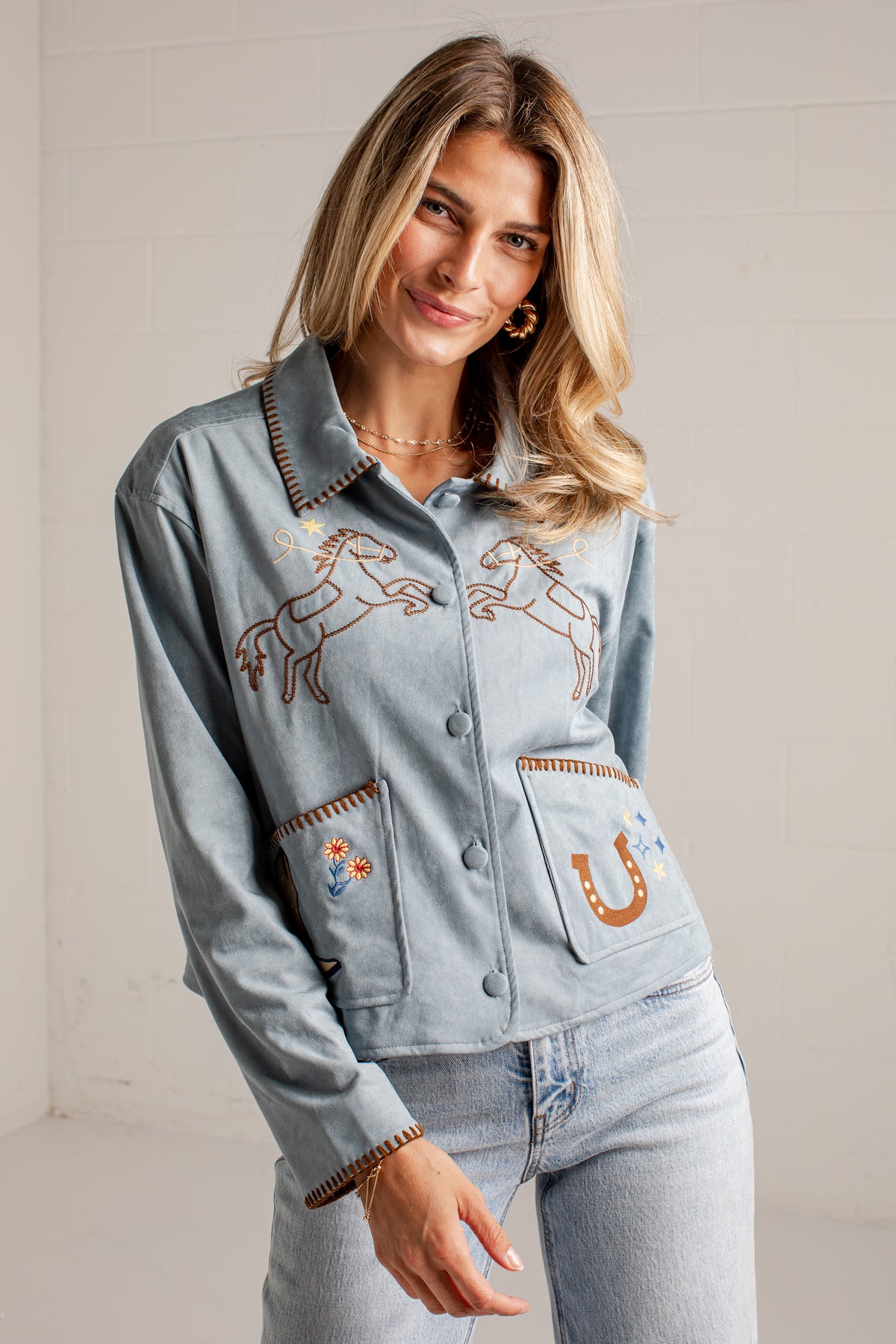 A close-up of a woman wearing a light blue suede jacket with western embroidery, including a horse design on the chest and a boot patch on the pocket.