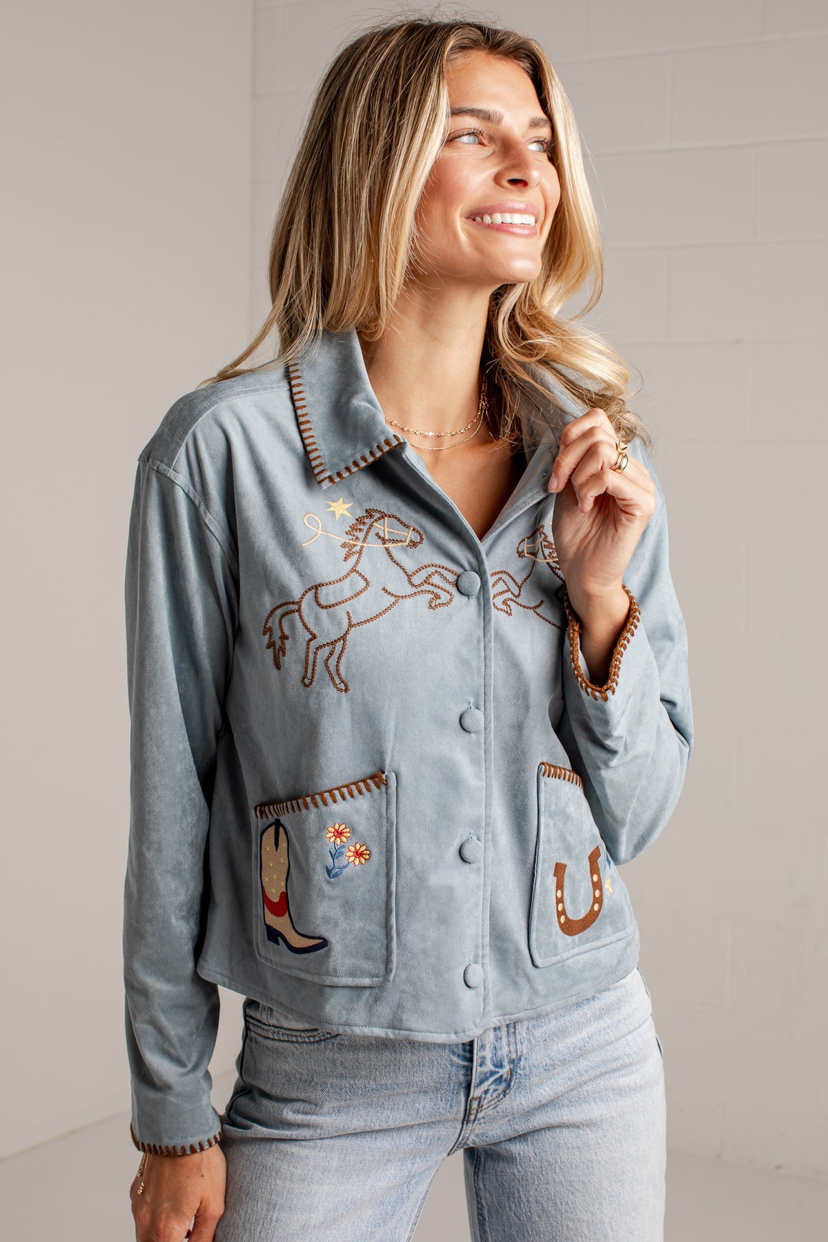 A close-up of a woman wearing a light blue suede jacket with western embroidery, including a horse design on the chest and a boot patch on the pocket.