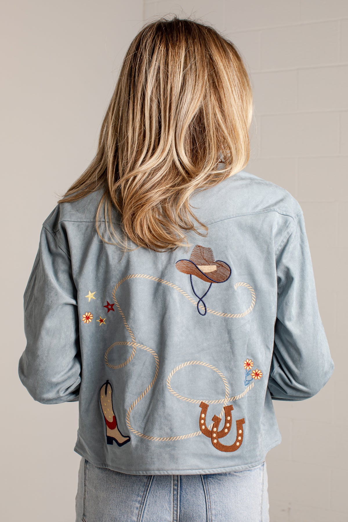 Back view of a person wearing a light blue denim jacket with embroidered western-themed designs including a cowboy hat, lasso, boots, stars, and horseshoes.