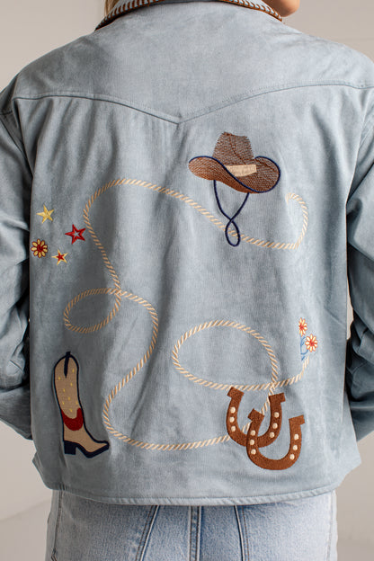 Back view of a person wearing a light blue denim jacket with embroidered western-themed designs including a cowboy hat, lasso, boots, stars, and horseshoes.