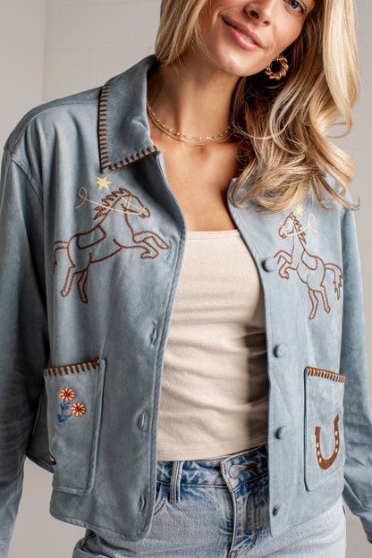 A close-up of a woman wearing a light blue suede jacket with western embroidery, including a horse design on the chest and a boot patch on the pocket.
