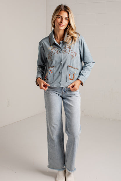 A woman wearing a light blue suede jacket with western embroidery, including a horse design on the chest and a boot patch on the pocket.