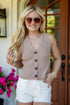 A woman wearing a sleeveless ribbed knit vest in a taupe-beige color with dark brown buttons, paired with white denim shorts, standing in front of a home entrance.