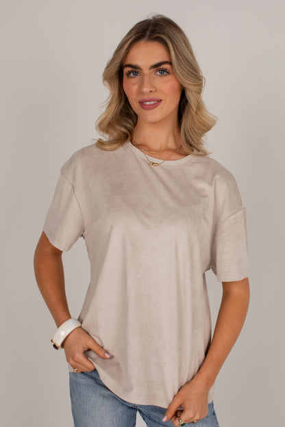 A woman wearing a light beige short-sleeve crewneck top and blue jeans, standing against a neutral light gray background.