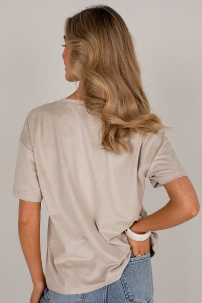 A woman with long blonde hair is shown from behind wearing a light beige short-sleeve t-shirt and blue jeans against a solid light gray background.