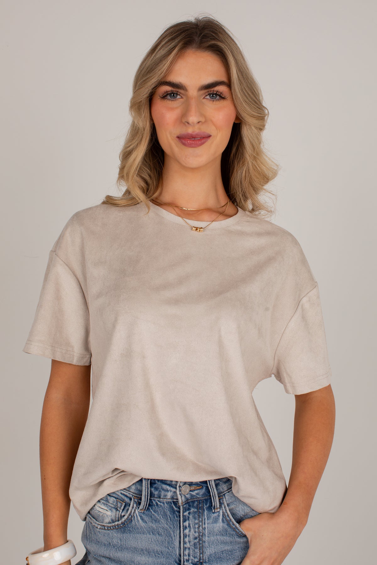 A woman wearing a light beige, short-sleeve crewneck t-shirt and blue jeans, standing against a neutral light gray background.
