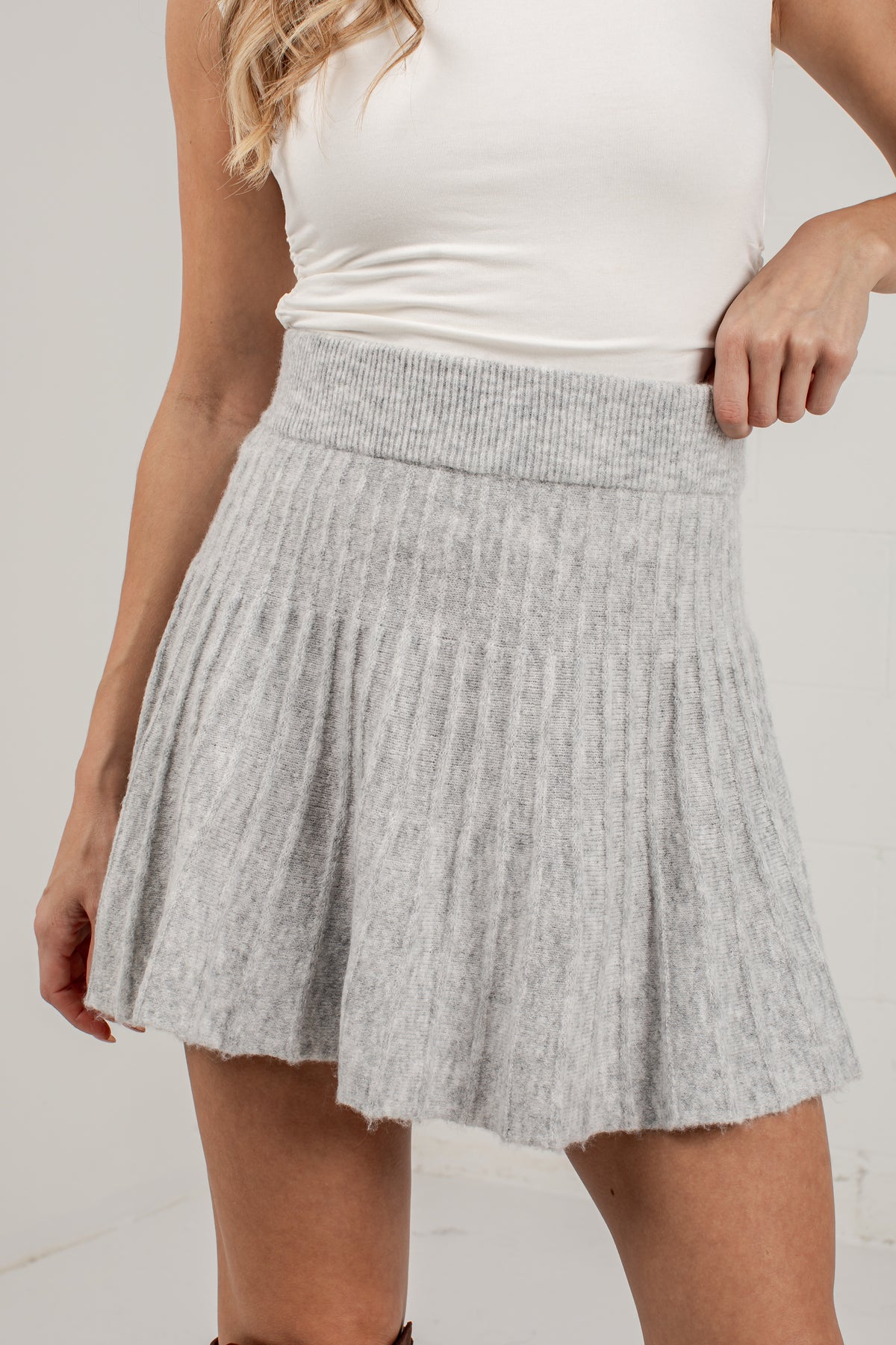 A woman wearing a light gray ribbed knit pleated mini skirt with a matching elastic waistband, paired with a white sleeveless top.