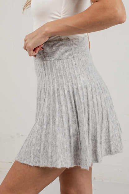 A light gray ribbed knit pleated mini skirt worn by a model, paired with a white top, against a white brick wall background.