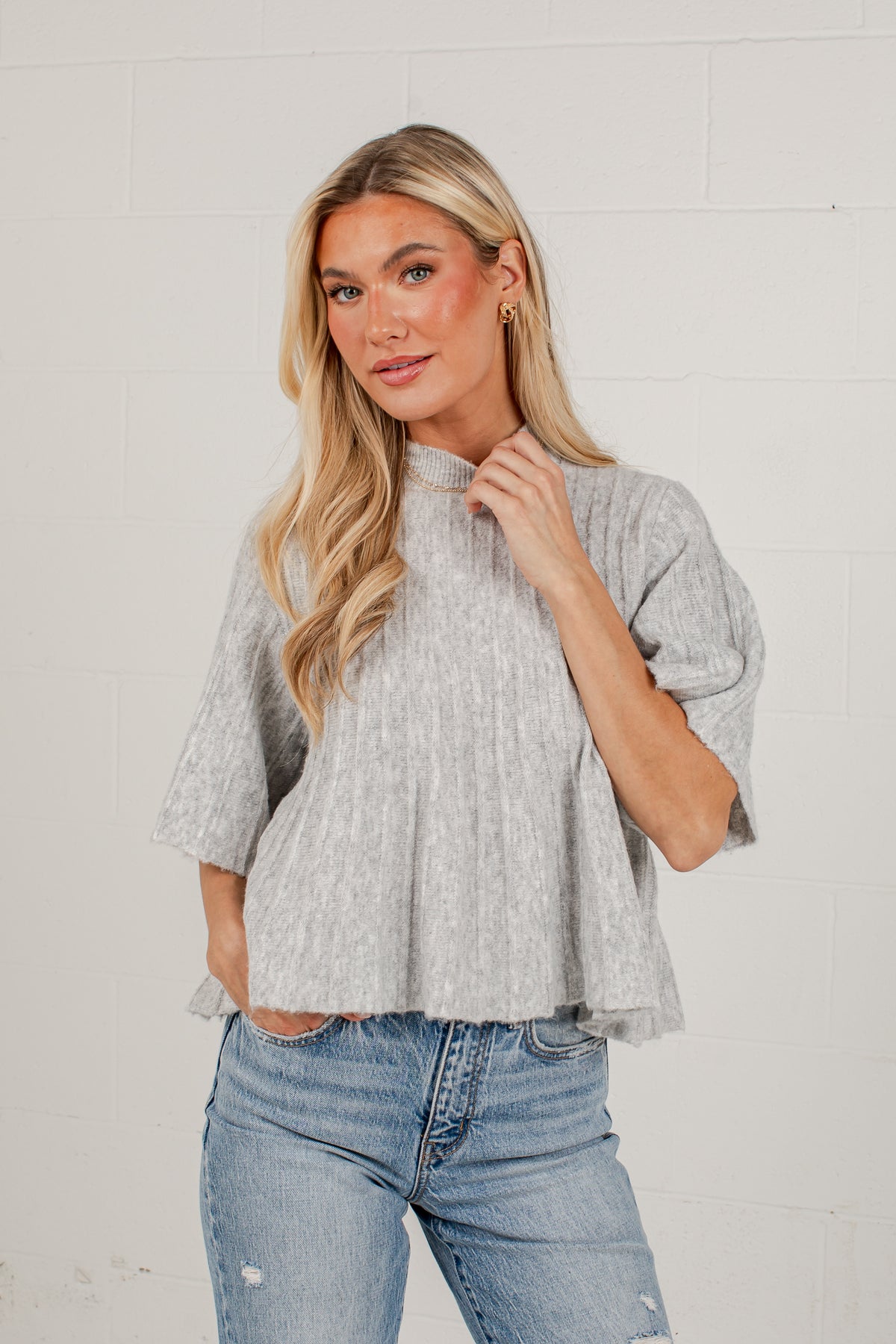A blonde woman wearing a light gray textured short-sleeve top with a high mock neck and blue jeans, standing against a white brick wall.