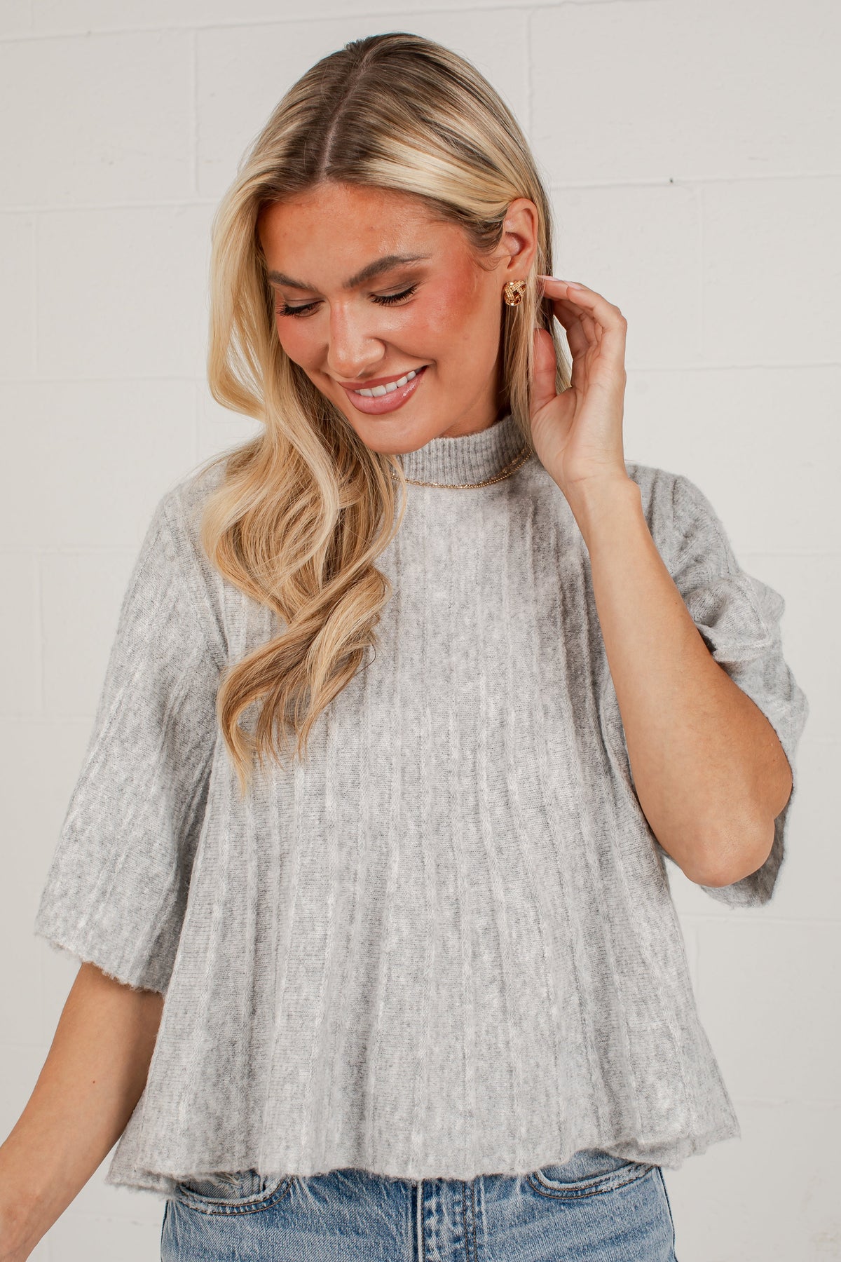 A woman wearing a light gray heathered short-sleeve mock neck top with a textured crinkle fabric, paired with blue jeans, standing against a white brick wall.