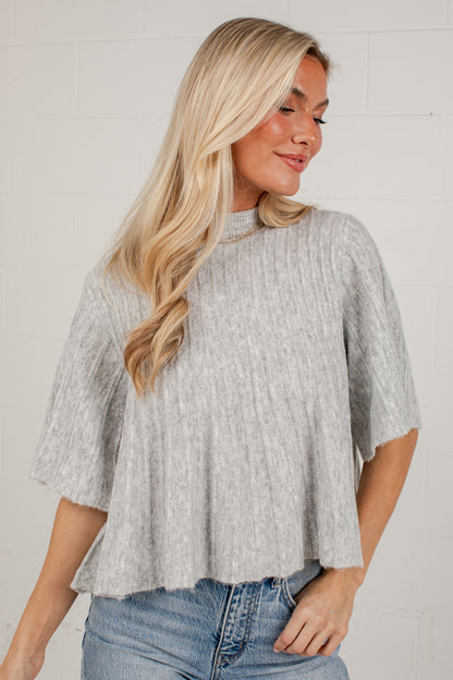 A woman wearing a light gray ribbed knit short-sleeve top with a high mock neck and a flared hem, paired with blue jeans, standing against a white brick wall.
