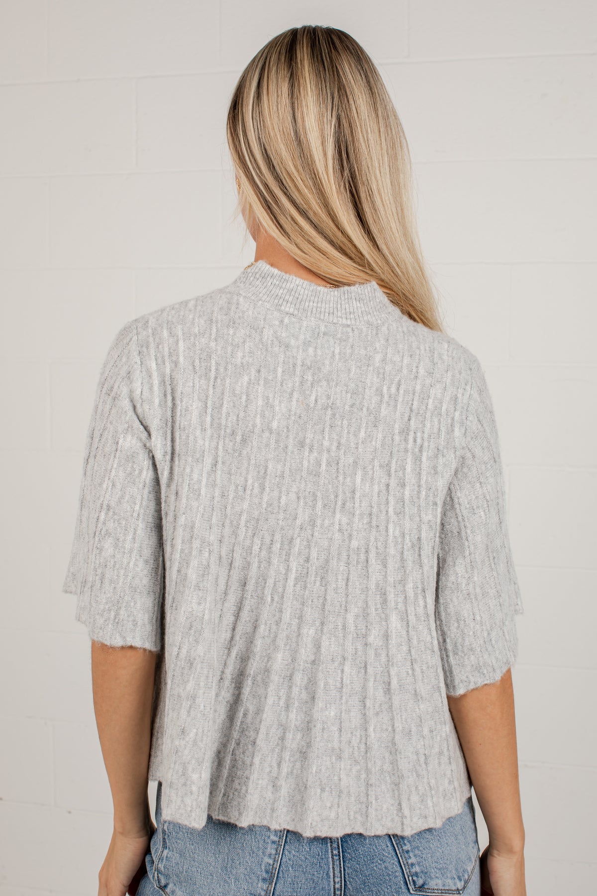 A woman viewed from behind wearing a light gray ribbed knit short-sleeve top and blue jeans against a white brick wall.