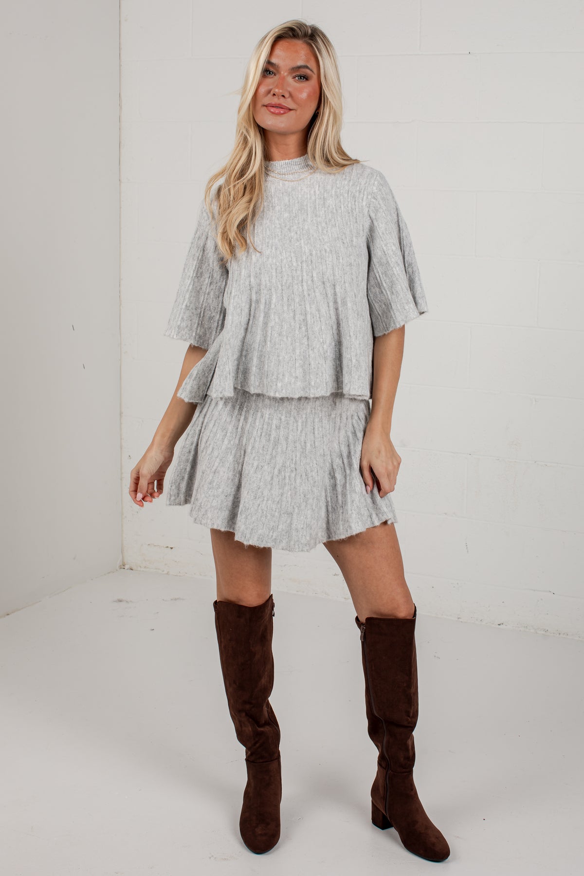 A woman modeling a light gray textured two-piece set consisting of a short-sleeve top and a pleated mini skirt, paired with brown knee-high boots, in a white studio setting.