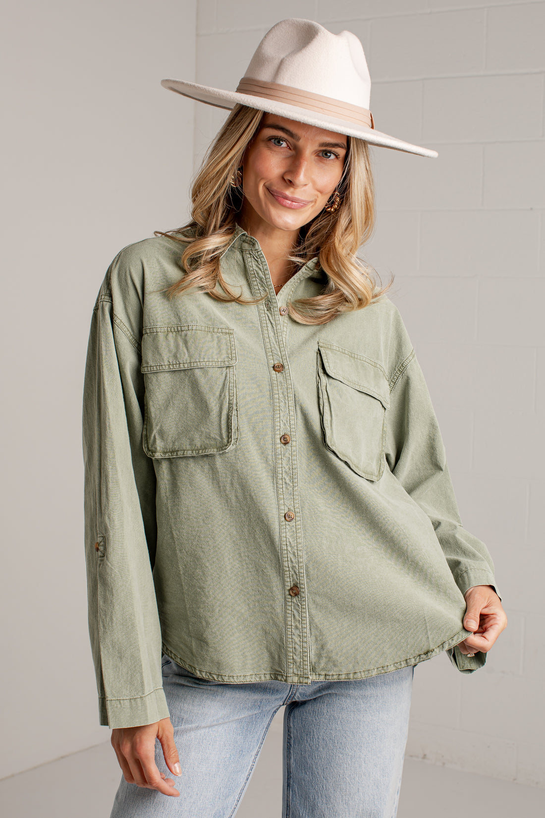 A woman wearing an oversized sage green button-up shirt with two chest pockets, paired with light-wash jeans and a wide-brimmed cream hat, standing against a light gray brick wall.