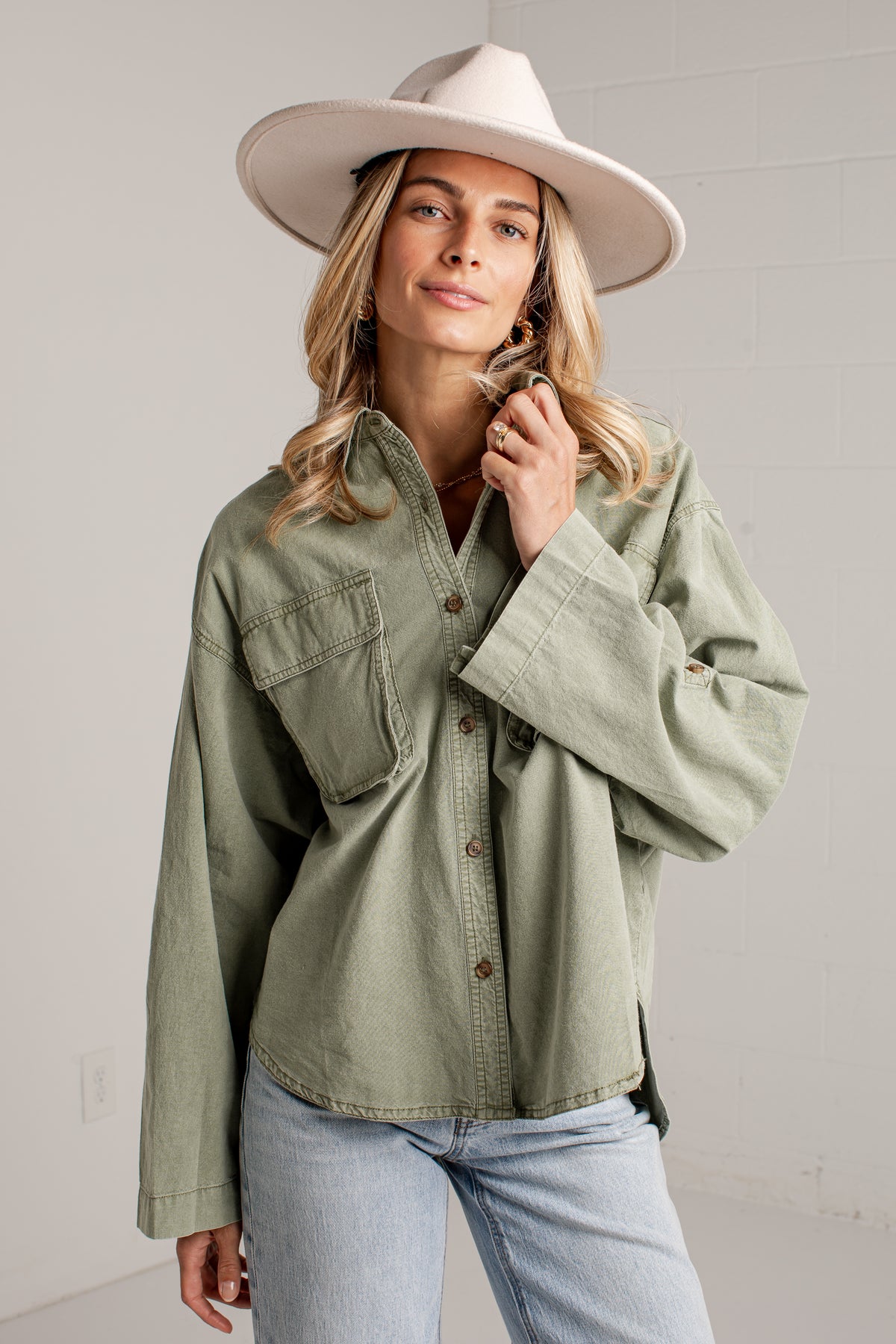 A woman wearing an oversized sage green button-up shirt with chest pockets, light-wash jeans, and a wide-brimmed cream hat, standing against a light gray wall.