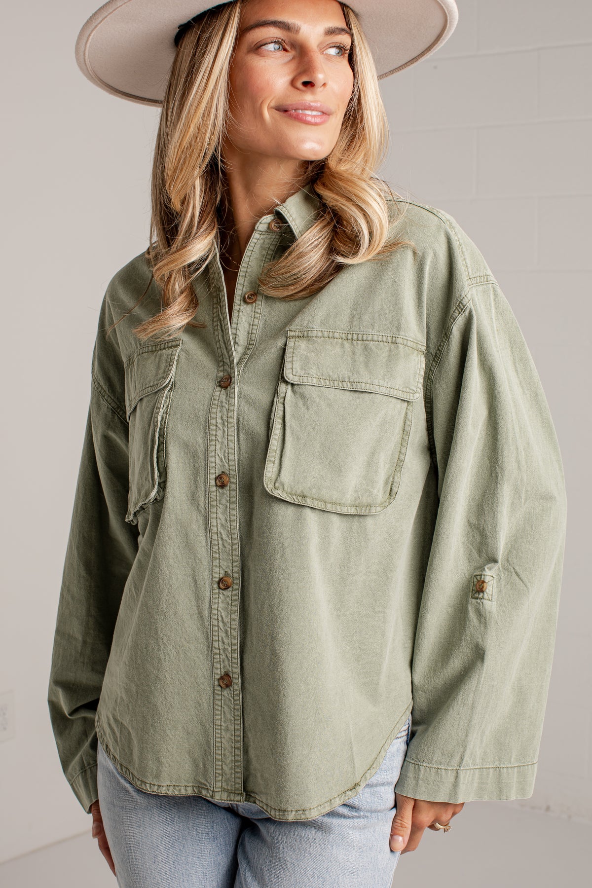 A woman wearing an oversized sage green button-up shirt with chest pockets, paired with light blue jeans and a wide-brimmed hat, photographed against a light gray wall.