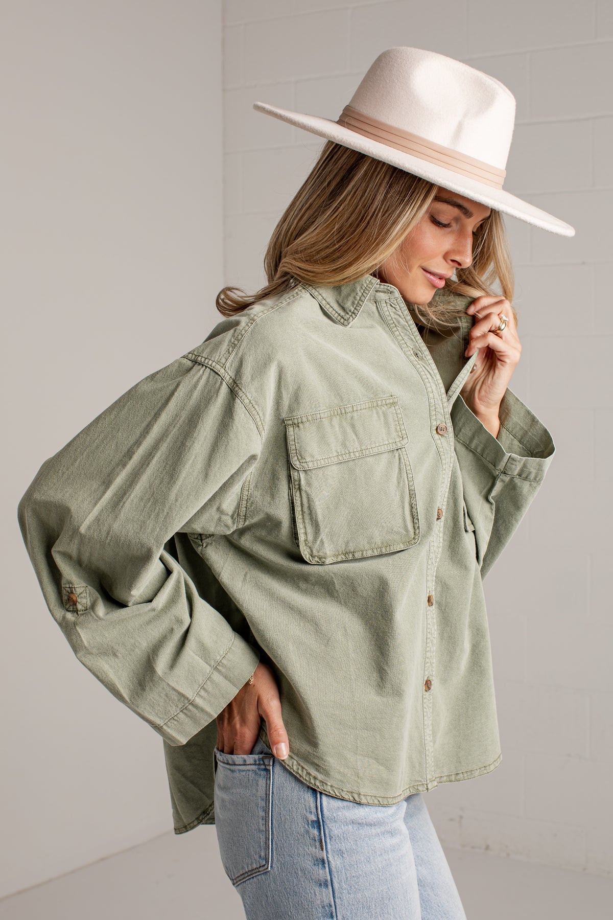 A woman wearing an oversized sage green button-up shirt with chest pockets, paired with light blue jeans and a wide-brimmed cream hat, posing against a light gray wall.