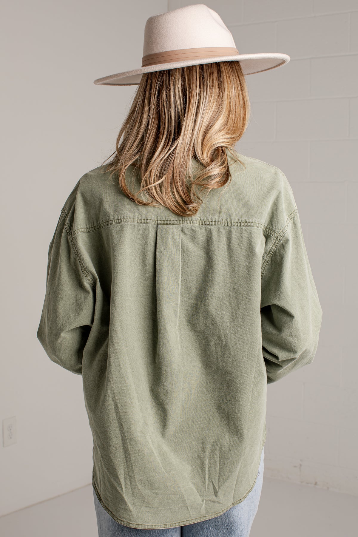 A woman shown from behind wearing an oversized sage green button-up shirt, light blue jeans, and a beige wide-brim fedora hat against a neutral light gray wall.