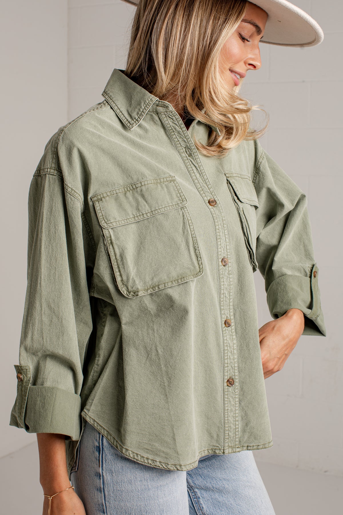 A woman wearing an oversized sage green button-up shirt with chest pockets and rolled sleeves, paired with light blue jeans and a white wide-brim hat, photographed against a light gray wall.