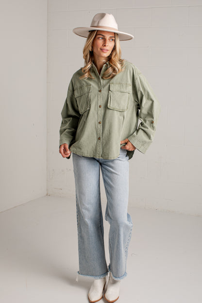 A woman modeling an oversized sage green button-up shirt with two chest pockets, paired with light-wash wide-leg jeans, white boots, and a beige wide-brim hat in a bright white studio setting.