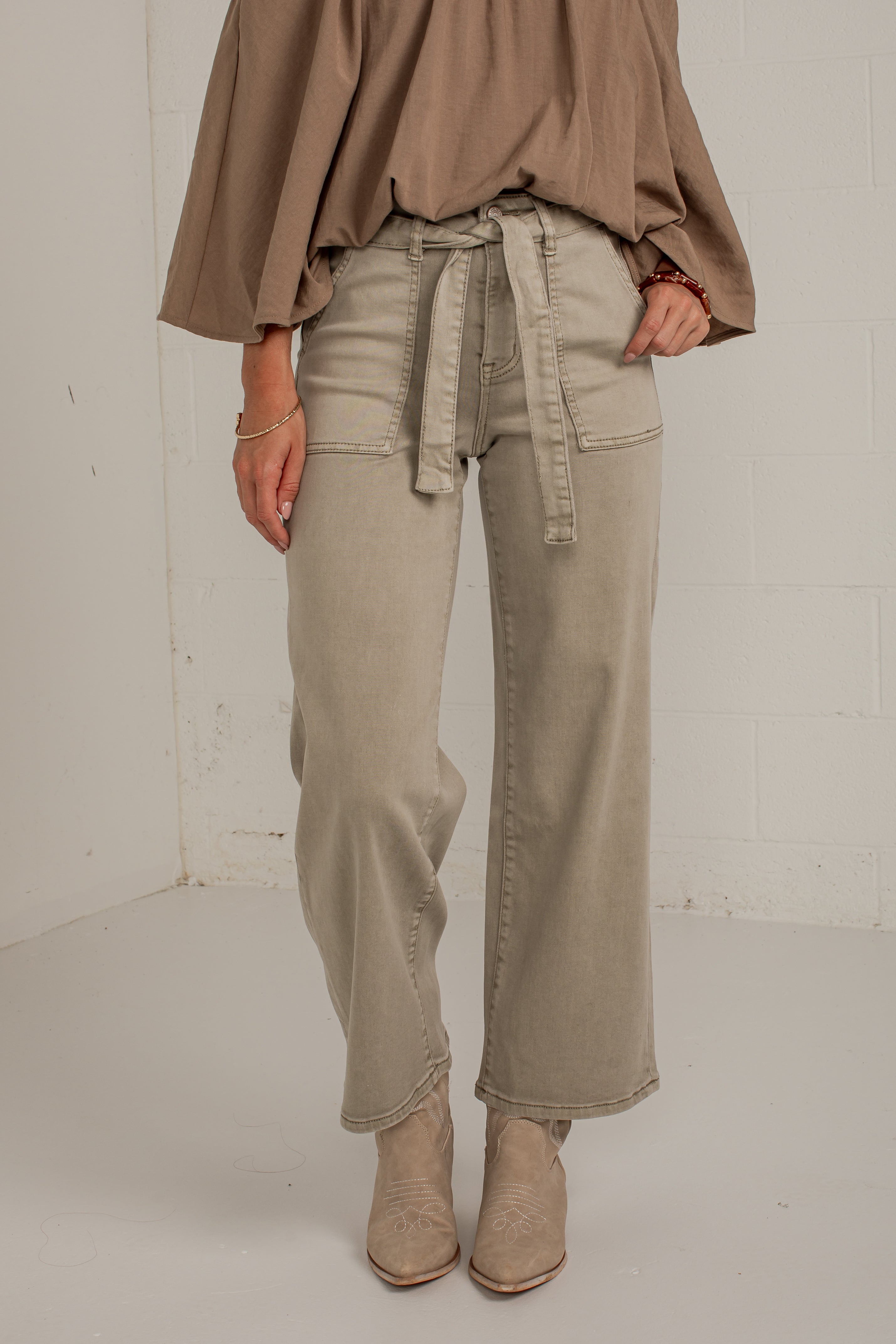A person wearing wide-leg beige jeans with a matching tie belt, paired with a taupe top and beige ankle boots, standing against a white brick wall.