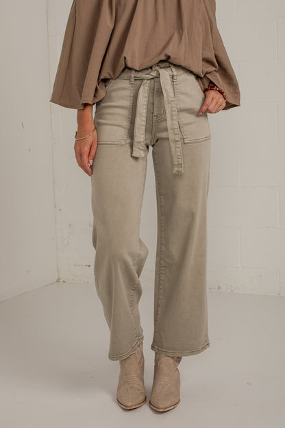 A person wearing wide-leg beige jeans with a matching tie belt, paired with a taupe top and beige ankle boots, standing against a white brick wall.