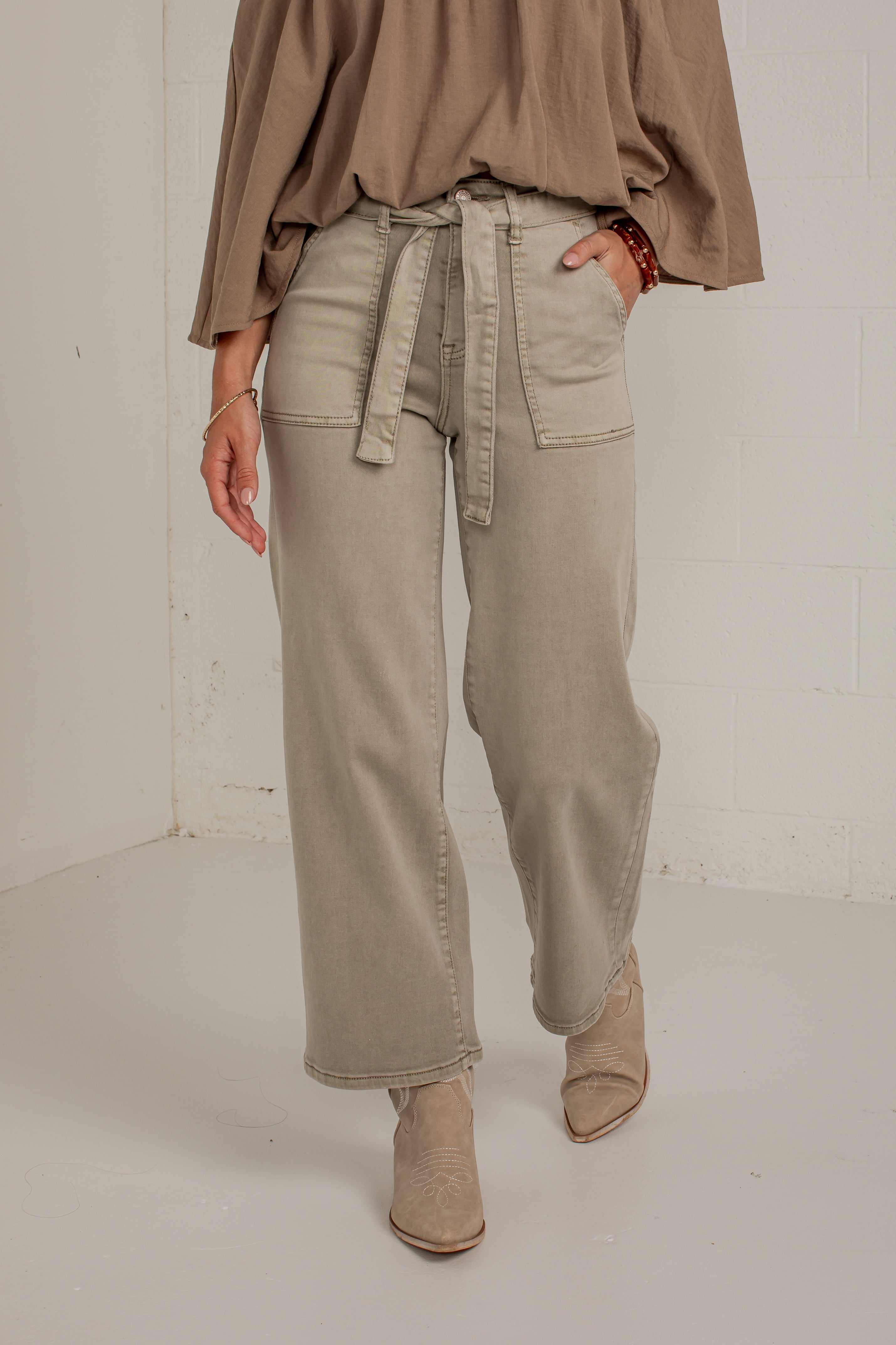 A model wearing a pair of wide-leg, light beige denim jeans with a matching fabric tie belt at the waist, styled with a taupe top and beige ankle boots.