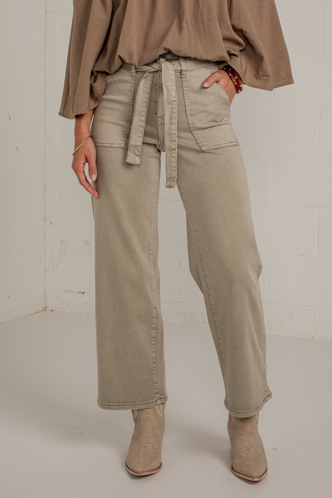 A model wearing a pair of wide-leg, straight-fit jeans in a light taupe-beige color. The jeans feature a matching fabric tie belt at the waist, side pockets, and a relaxed, straight-leg silhouette.