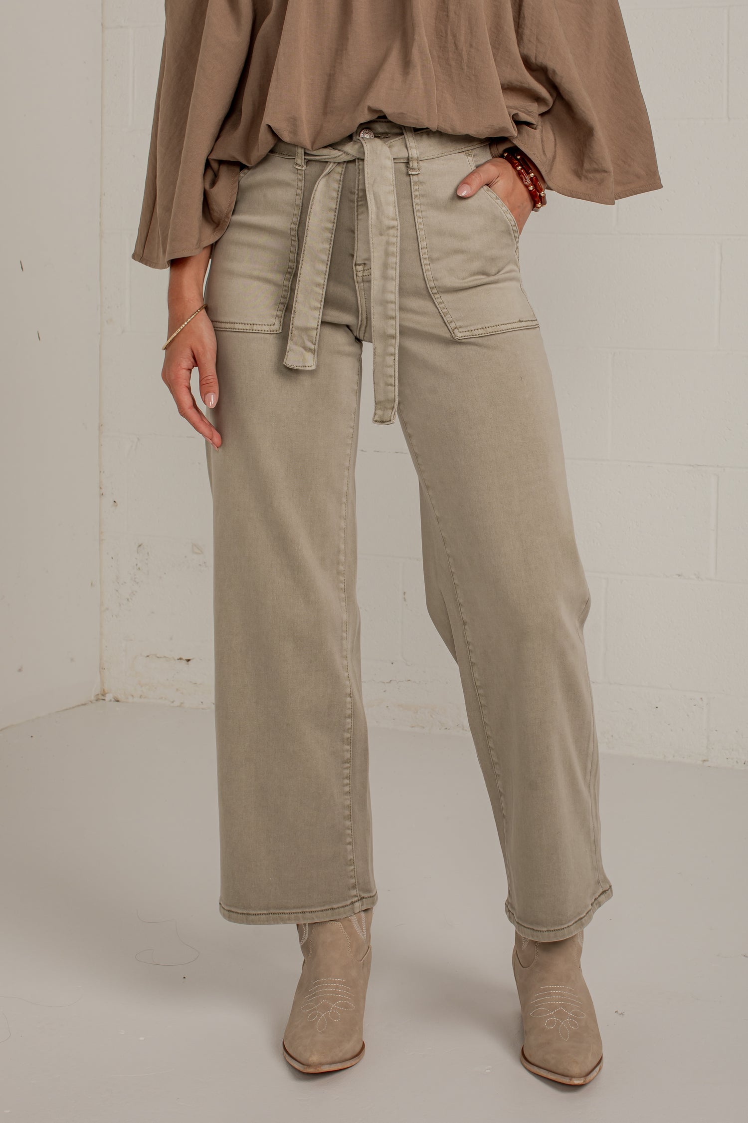 A model wearing a pair of wide-leg, straight-fit jeans in a light taupe-beige color. The jeans feature a matching fabric tie belt at the waist, side pockets, and a relaxed, straight-leg silhouette.