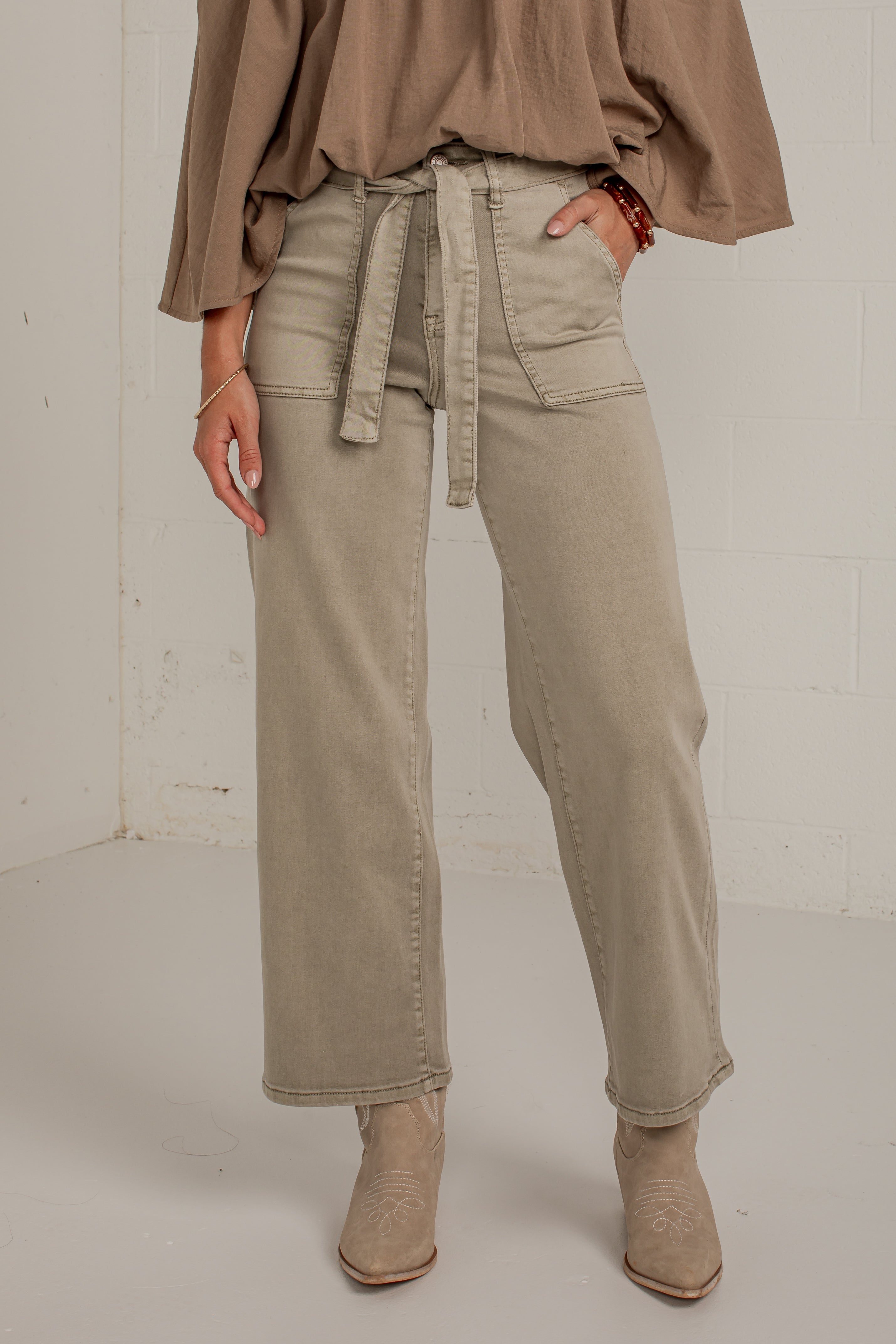 A model wearing a pair of wide-leg, straight-fit jeans in a light taupe-beige color. The jeans feature a matching fabric tie belt at the waist, side pockets, and a relaxed, straight-leg silhouette.