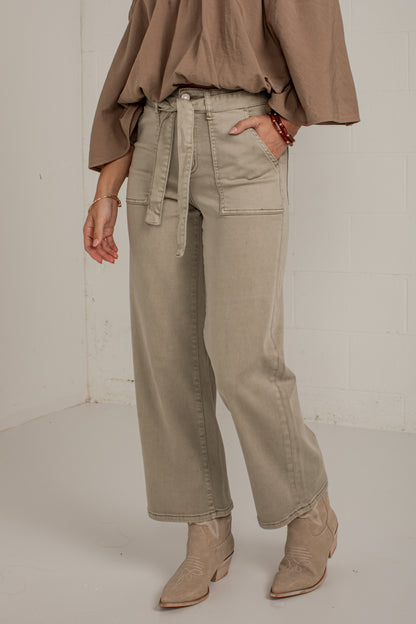 A person wearing light taupe wide-leg cargo pants with a matching tie belt, paired with a brown top and beige ankle boots, standing against a white studio wall.