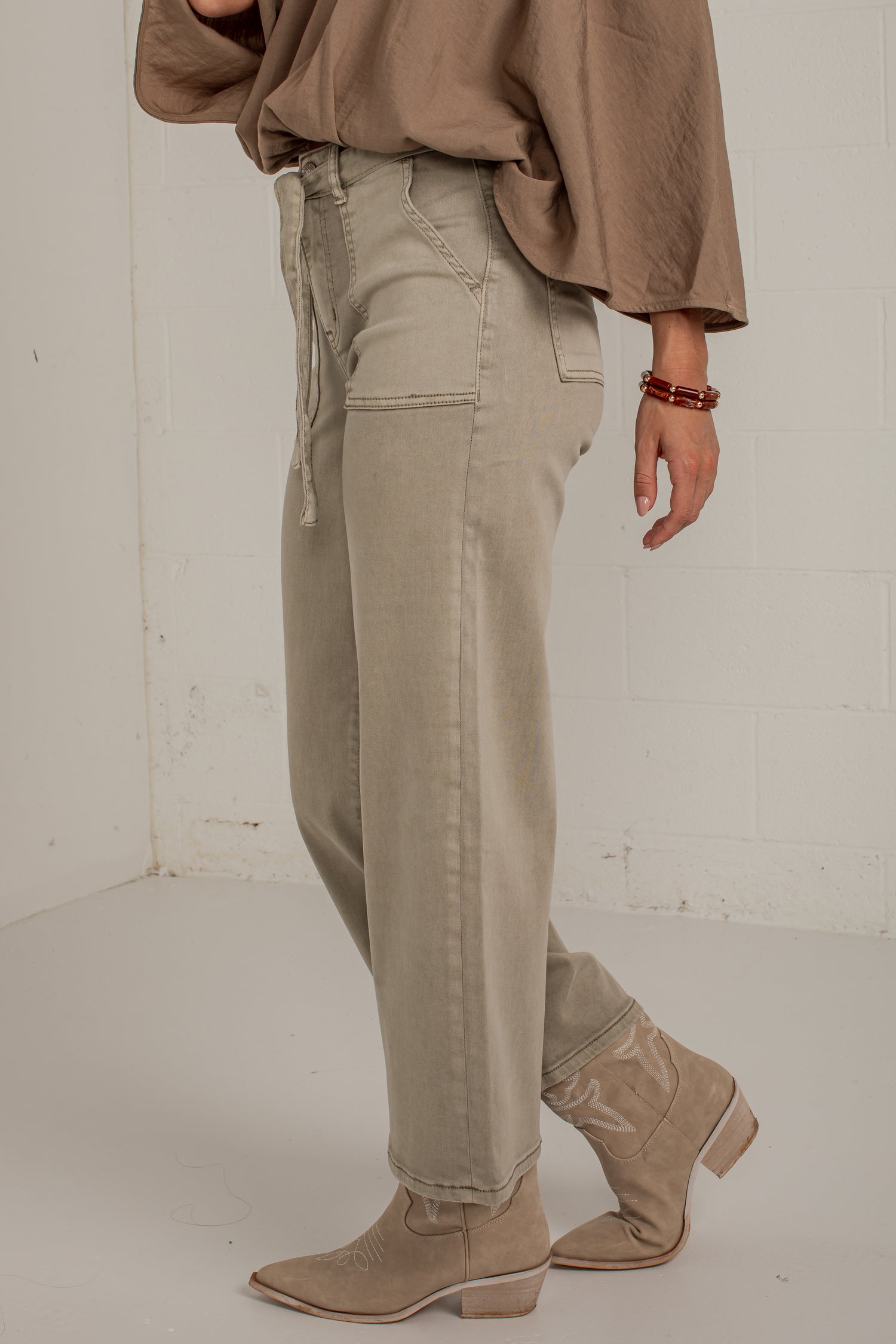 A person wearing light beige wide-leg jeans with a drawstring waist, paired with a taupe top and beige ankle boots, standing against a white brick wall.