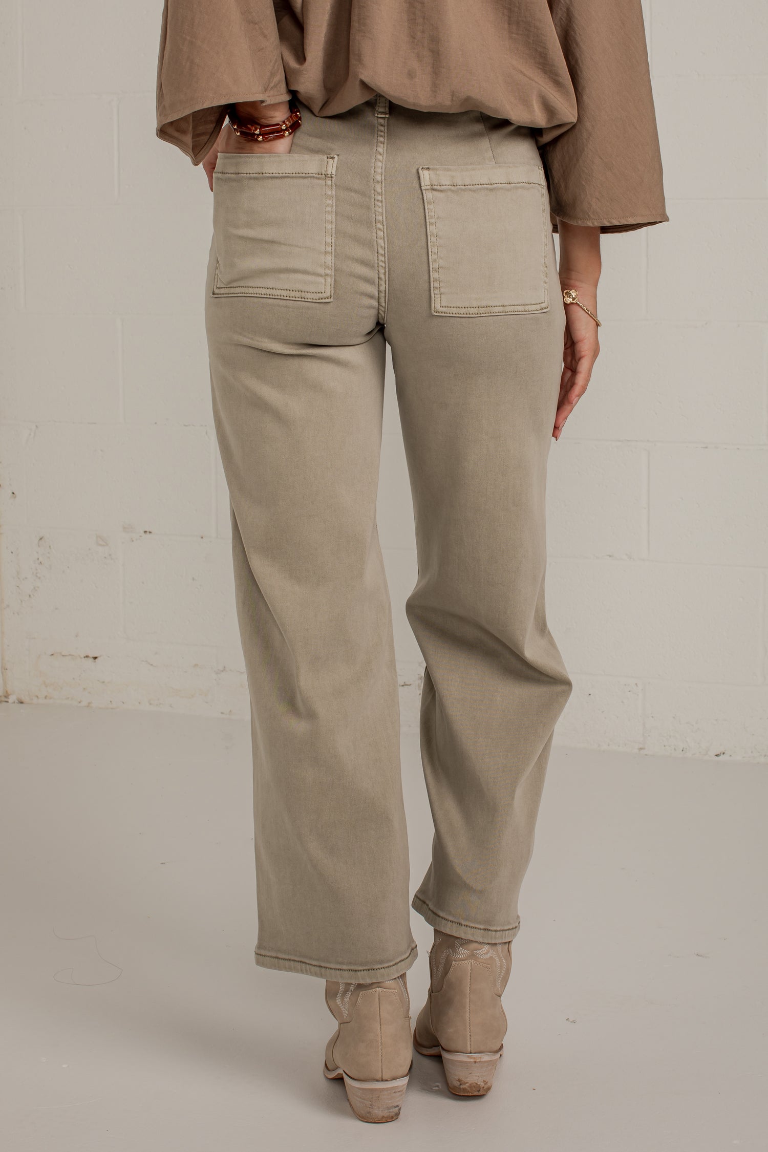 A person shown from the back wearing light beige wide-leg jeans with a brown top and beige ankle boots, standing against a white brick wall.
