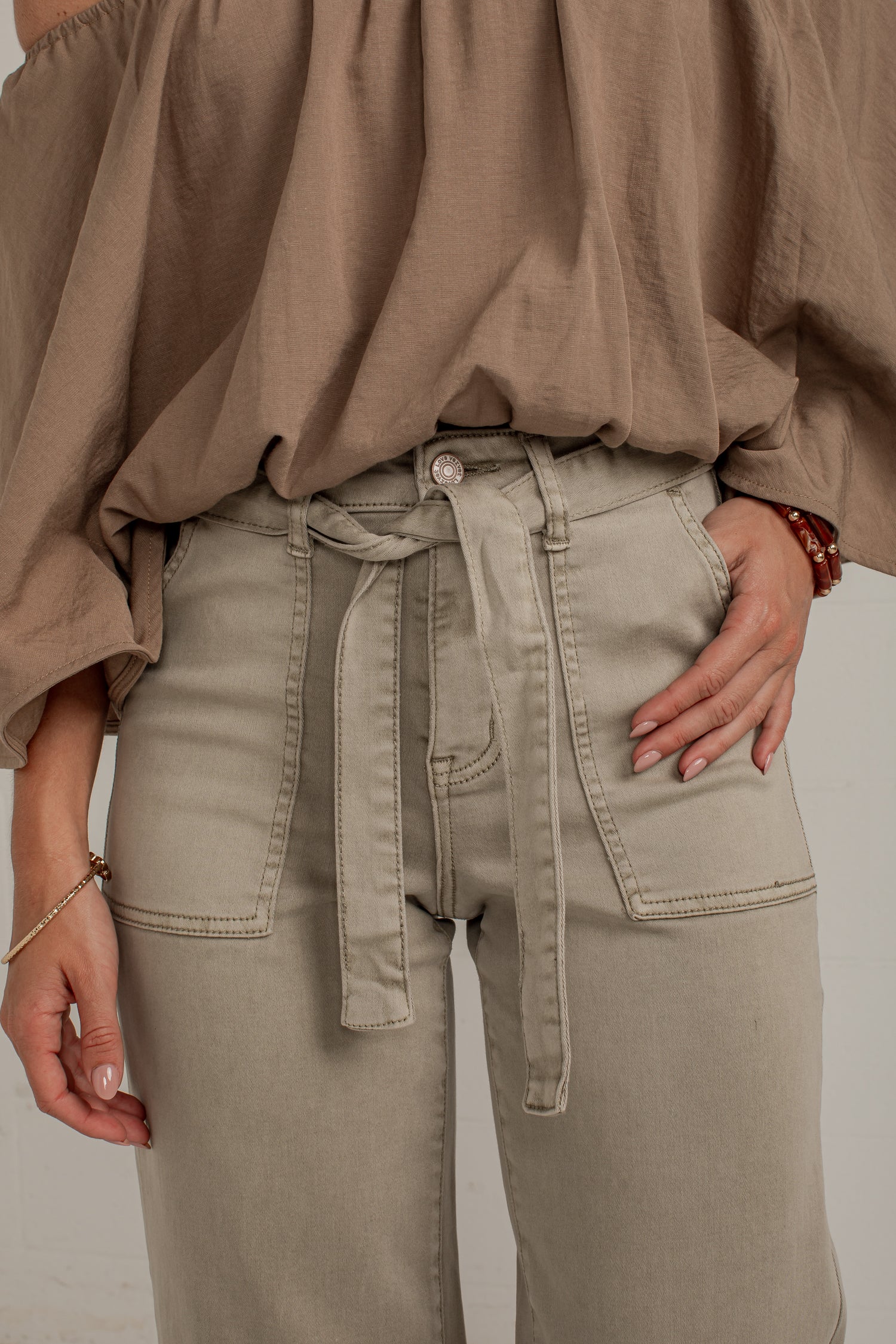 Close-up of a person wearing light beige high-waisted pants with a matching fabric tie belt and a taupe off-the-shoulder blouse against a plain light background.