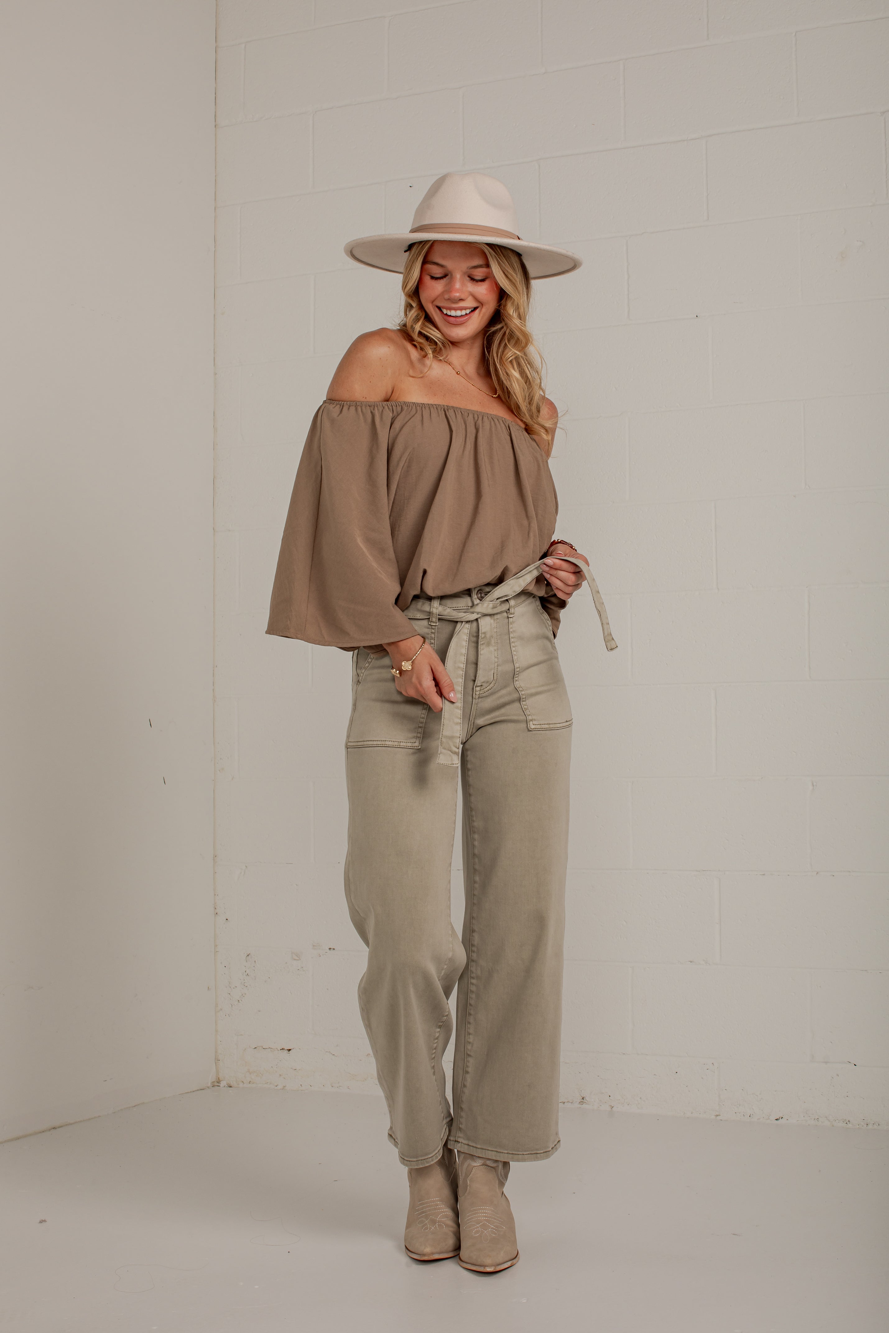 A woman modeling a taupe off-the-shoulder top with wide bell sleeves, paired with light beige wide-leg jeans, a matching hat, and beige ankle boots, standing against a white brick wall.