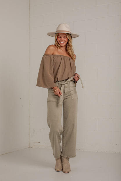 A woman modeling a taupe off-the-shoulder top with wide bell sleeves, paired with light beige wide-leg jeans, a matching hat, and beige ankle boots, standing against a white brick wall.