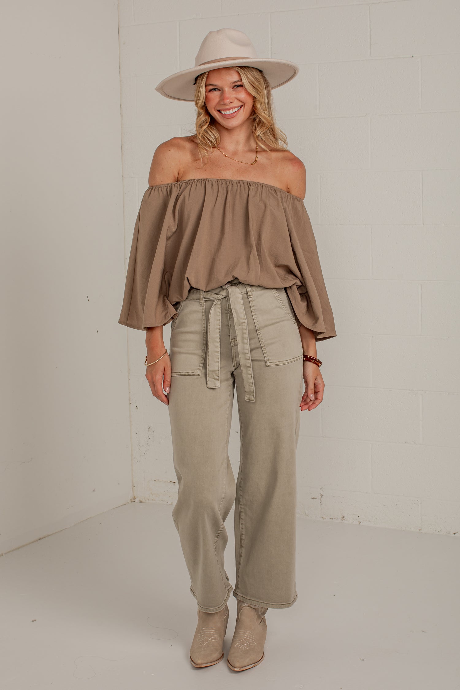 A woman modeling an off-the-shoulder taupe top, wide-leg khaki pants, beige ankle boots, and a beige wide-brim hat against a white brick wall background.