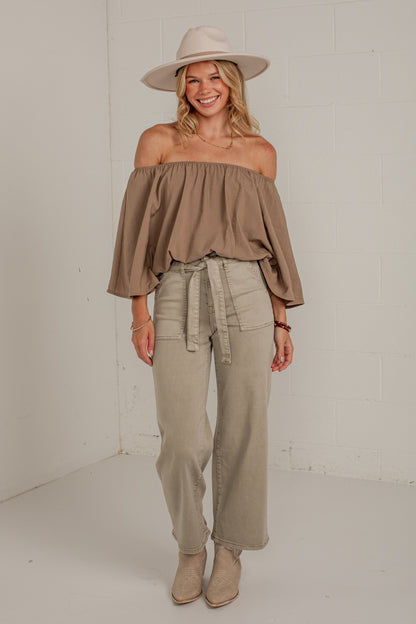 A woman modeling an off-the-shoulder taupe top, wide-leg khaki pants, beige ankle boots, and a beige wide-brim hat against a white brick wall background.