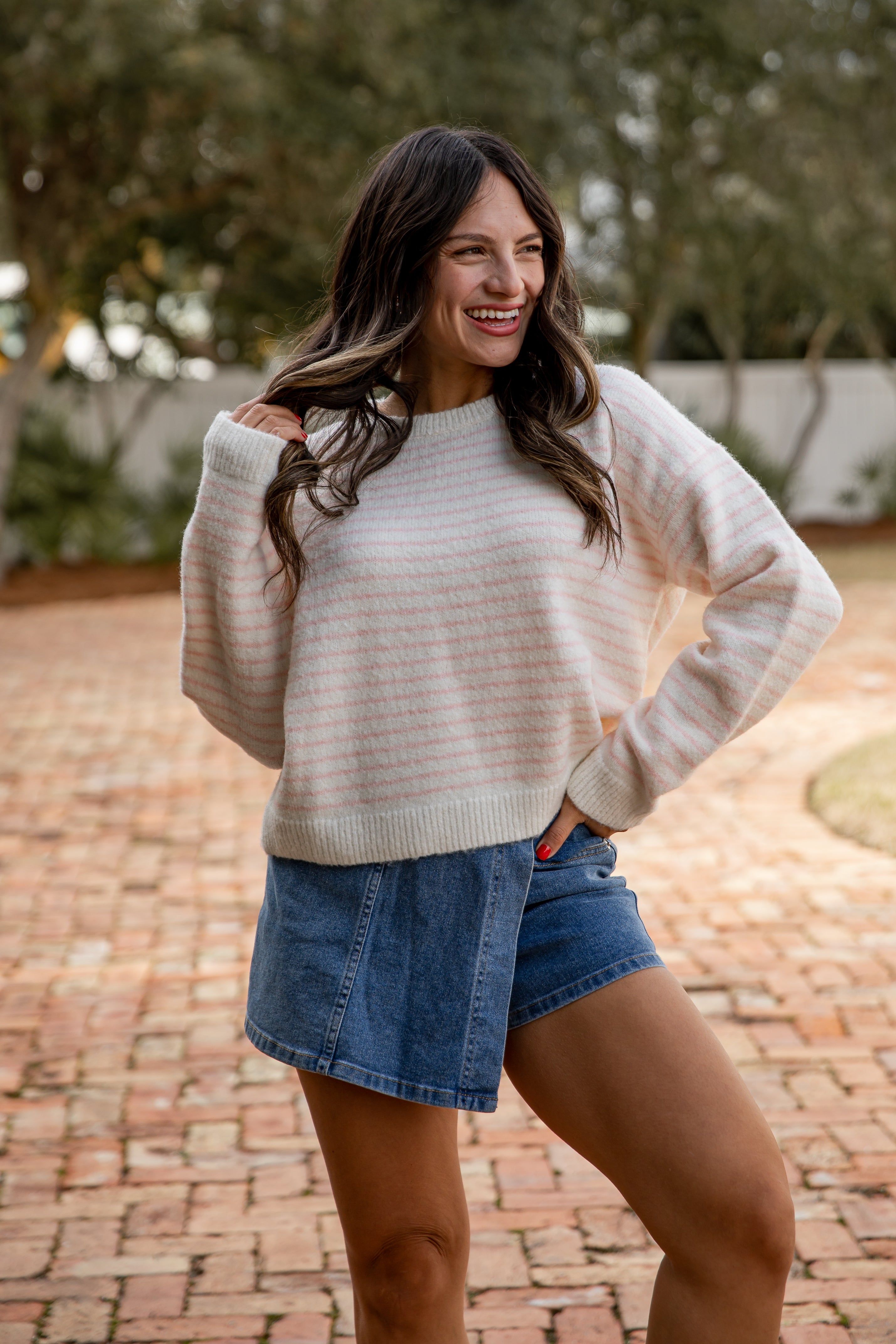 Woman wearing a beige sweater and denim shorts standing on a brick path with trees in the background