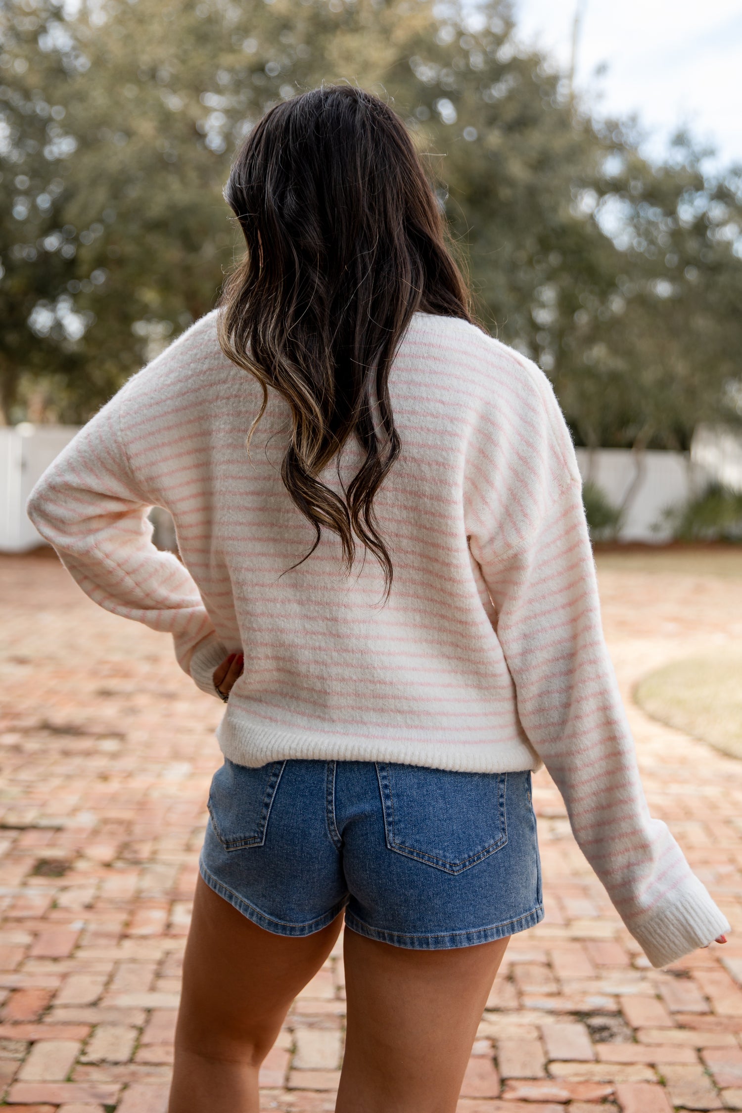 Person wearing a beige sweater and denim shorts standing outdoors on a brick path with trees in the background.