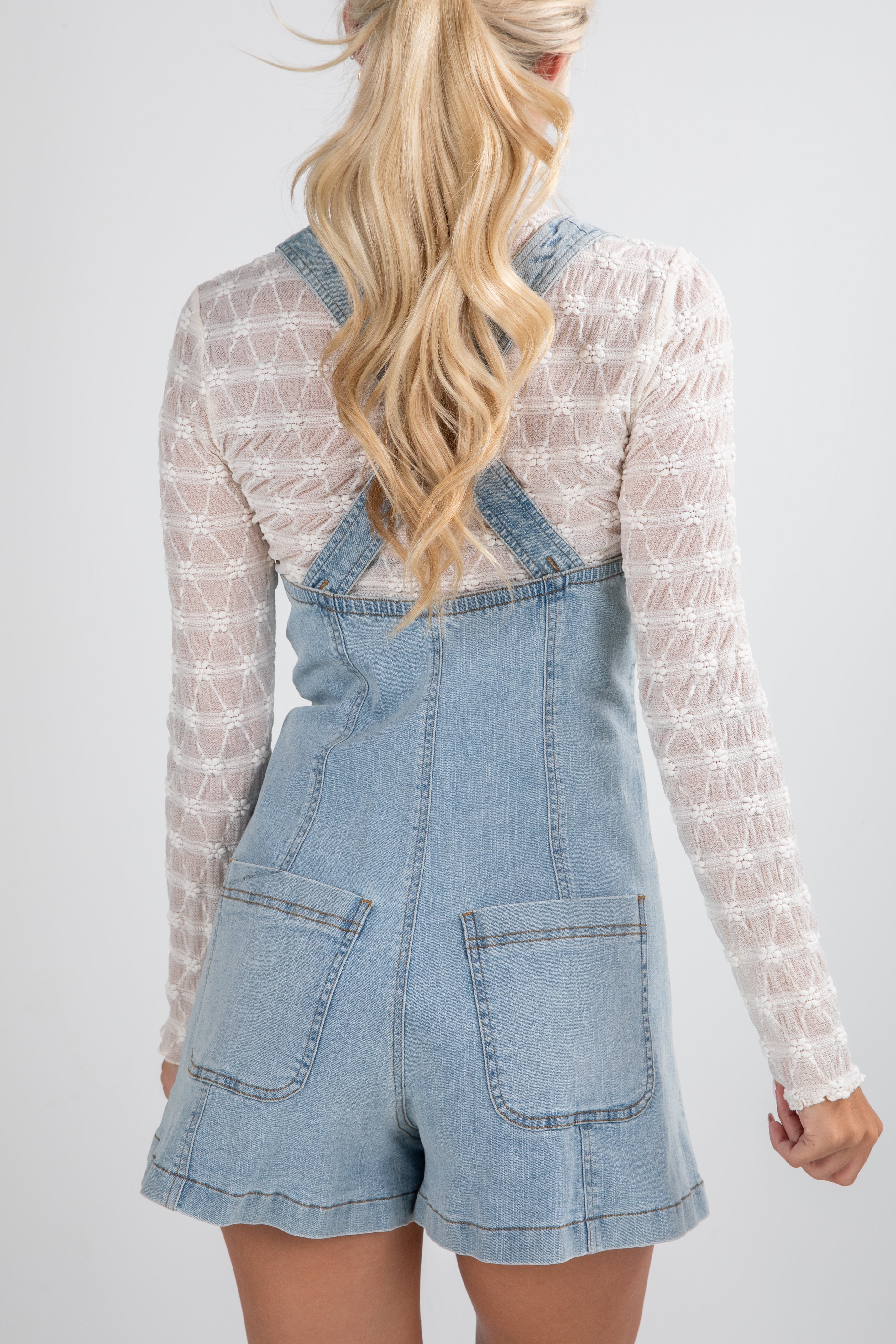 Person wearing a denim overall dress with a white lace top on a plain background