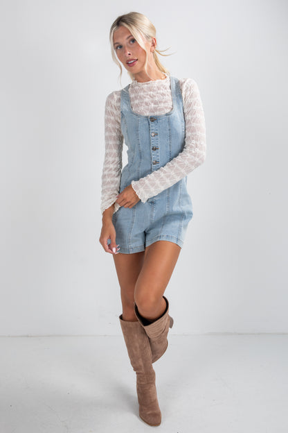Woman wearing a denim pinafore dress with a white lace top and brown boots on a white background